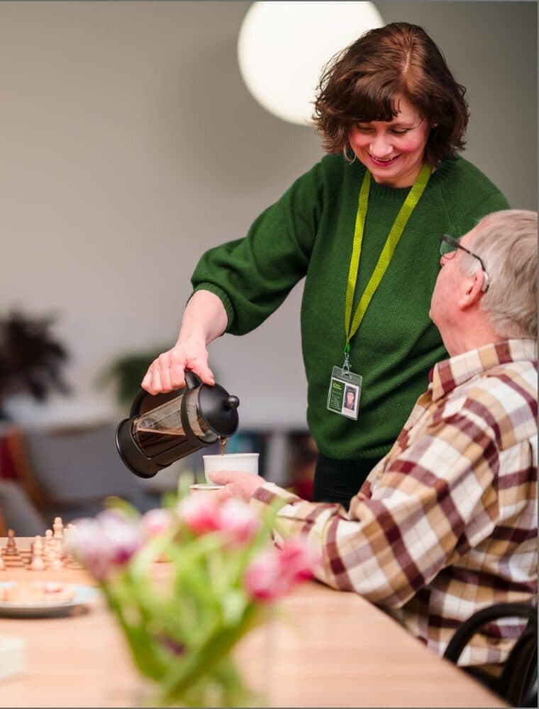 A woman with a name tag smiling while holding a dog, seated across from an older man in a living room. - Home Instead