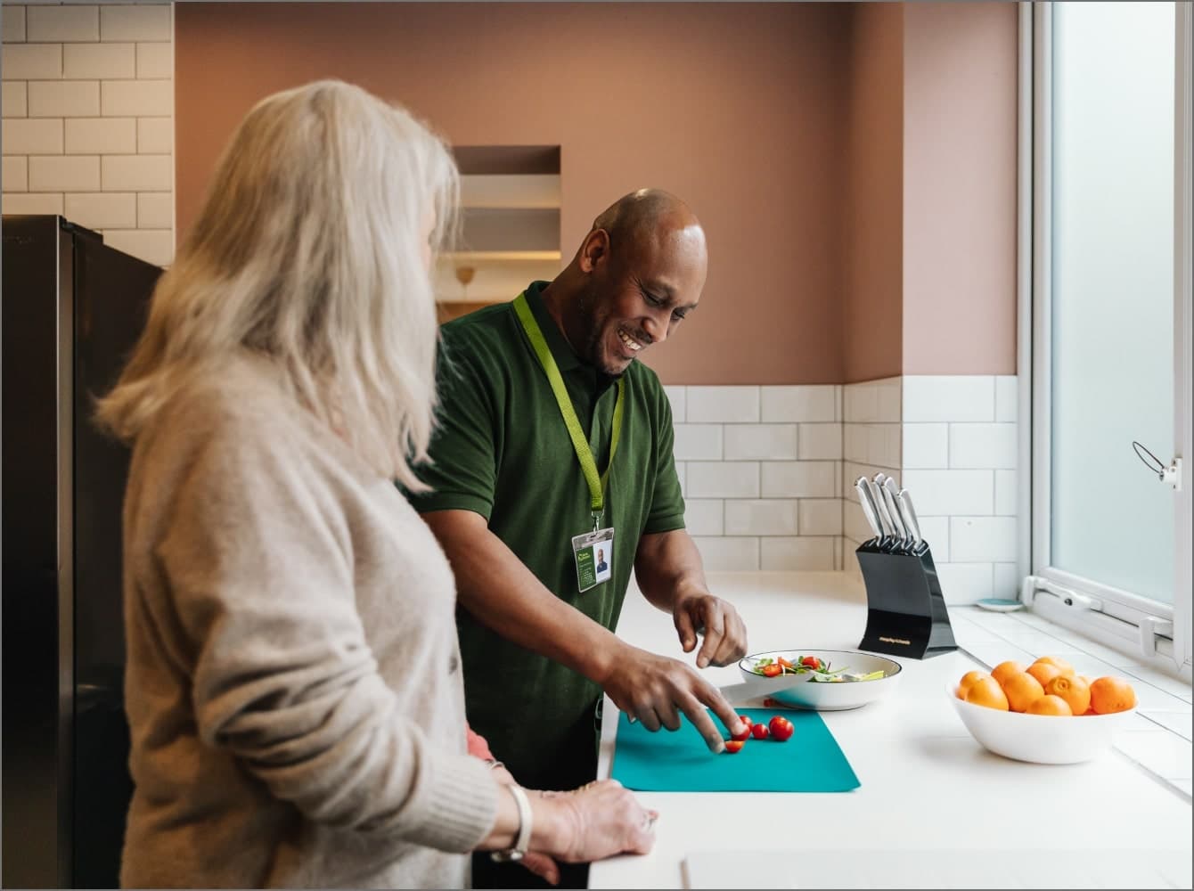 Two people in a kitchen, one slicing tomatoes on a cutting board, both smiling and interacting. Companionship Care at Home Instead Taunton & West Somerset
