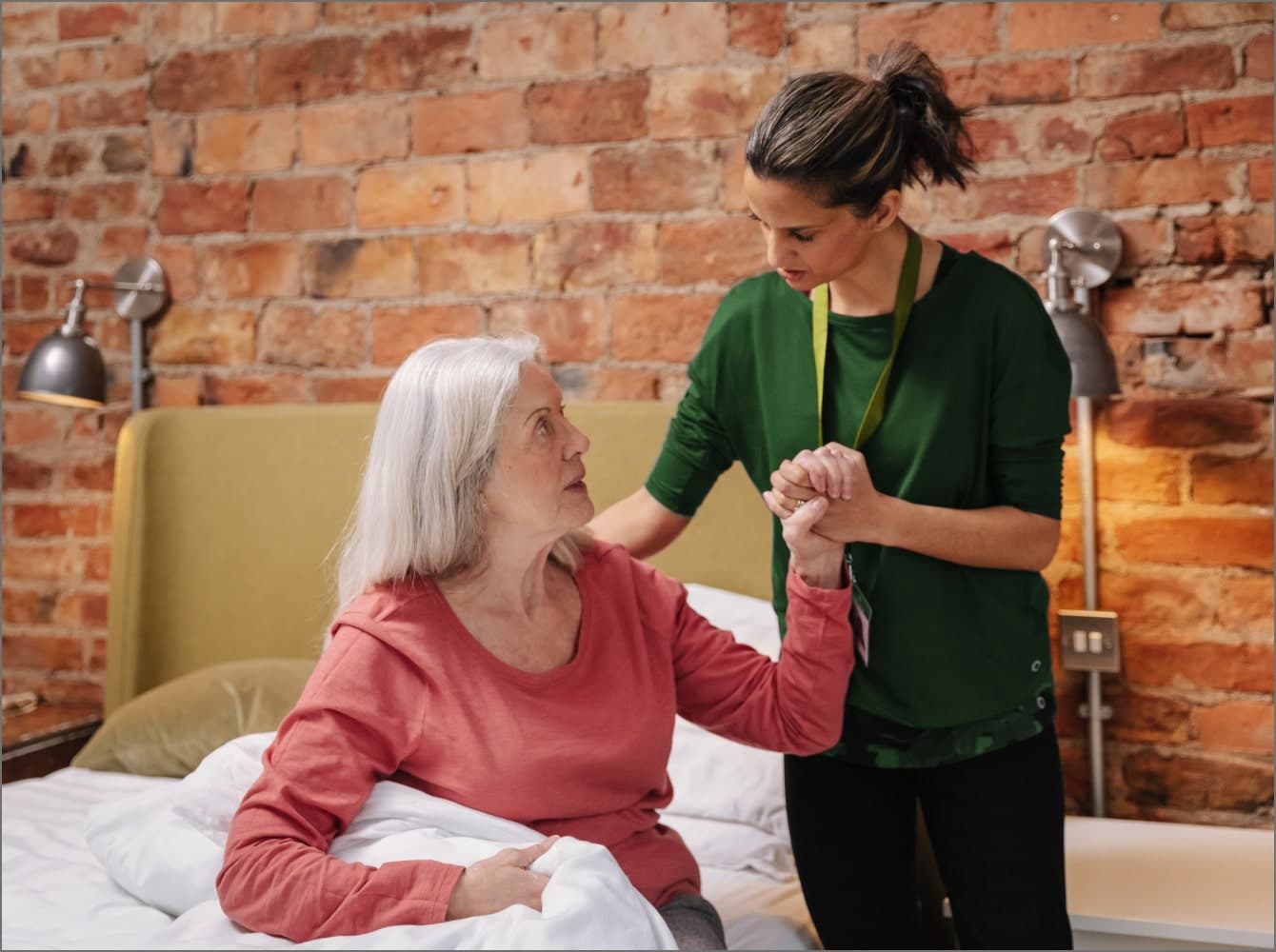 A caregiver helps an elderly woman sit up in bed in a room with brick walls. Companionship Care at Home Instead Taunton & West Somerset