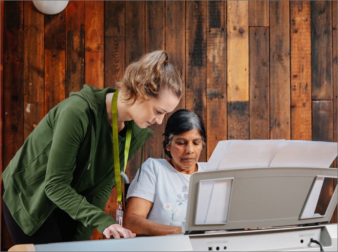 Two women playing the piano, focused on sheet music, against a wooden wall background. Companionship Care at Home Instead Taunton & West Somerset