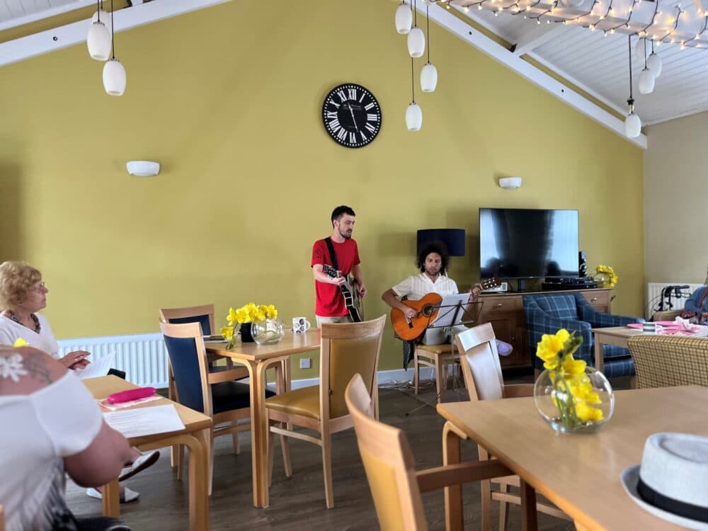 Two musicians with guitars perform in a room decorated with flowers and a clock on the wall, watched by seated people. - Home Instead