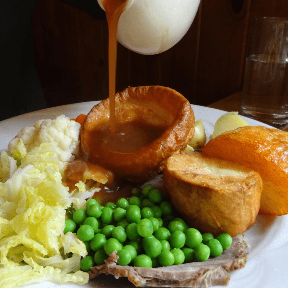A plate with roast beef, Yorkshire pudding, peas, potatoes, cabbage, and squash is being topped with gravy. - Home Instead