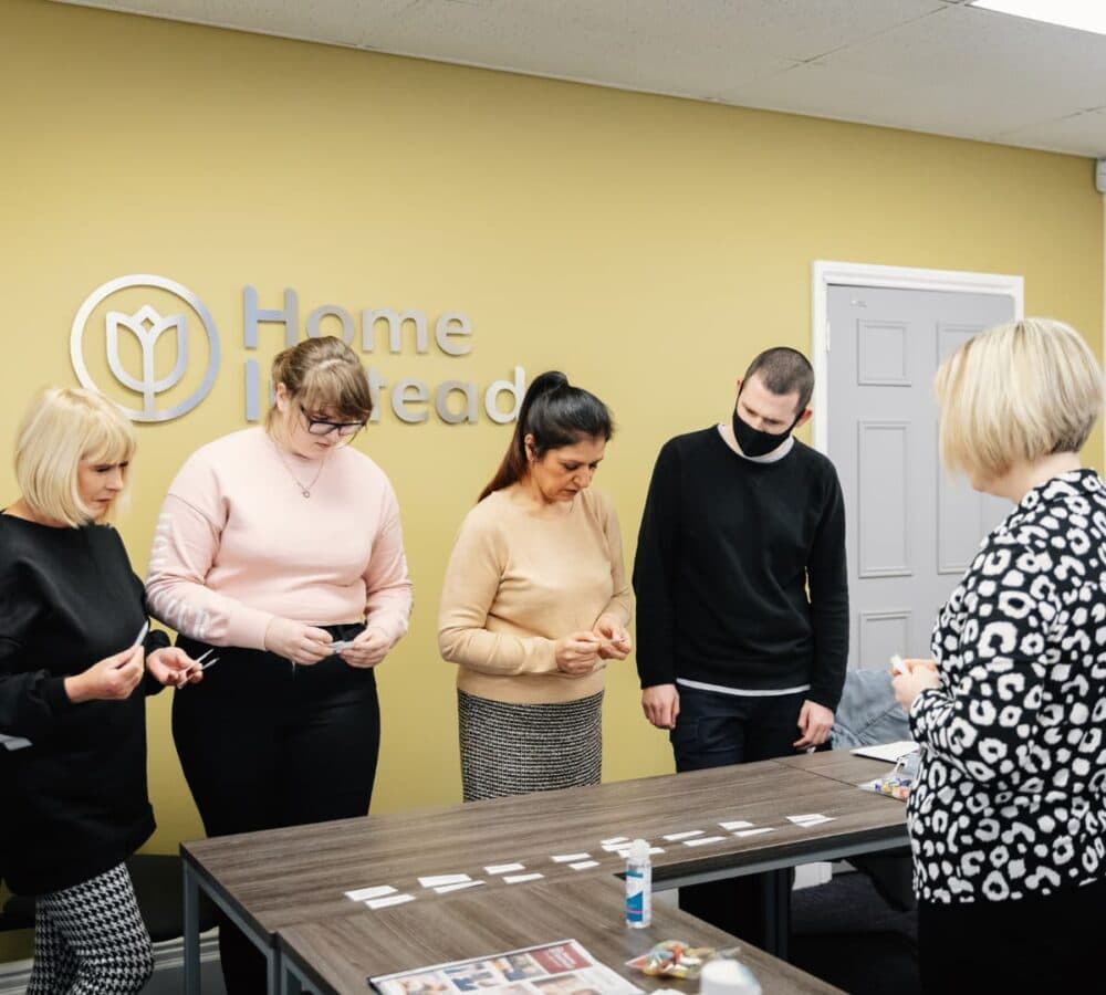 A man wearing black and a mask and a woman with long hair, wearing beige and another woman wearing pink with eyeglasses and another woman wearing black with short blonde hair inside a room looking at the table with a beige background and with Home Instead logo