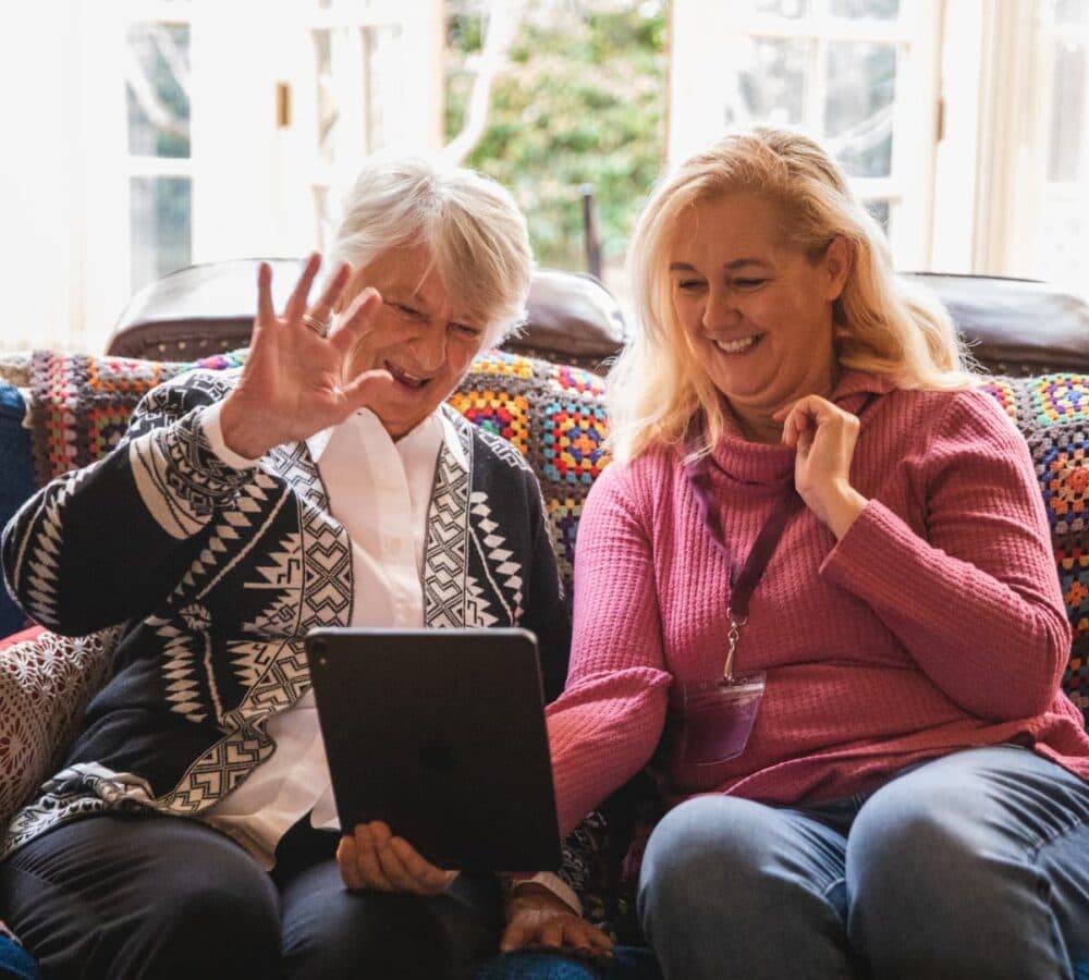 A woman wearing pink with long blonde hair and wearing an ID with an older woman with white hair and white top looking at an IPAD while smiling and waving and while sitting on a couch with a window background inside a house