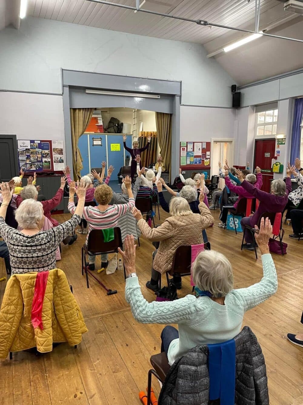 A group of seniors seated on chairs in a hall, raising their hands during an exercise session. - Home Instead