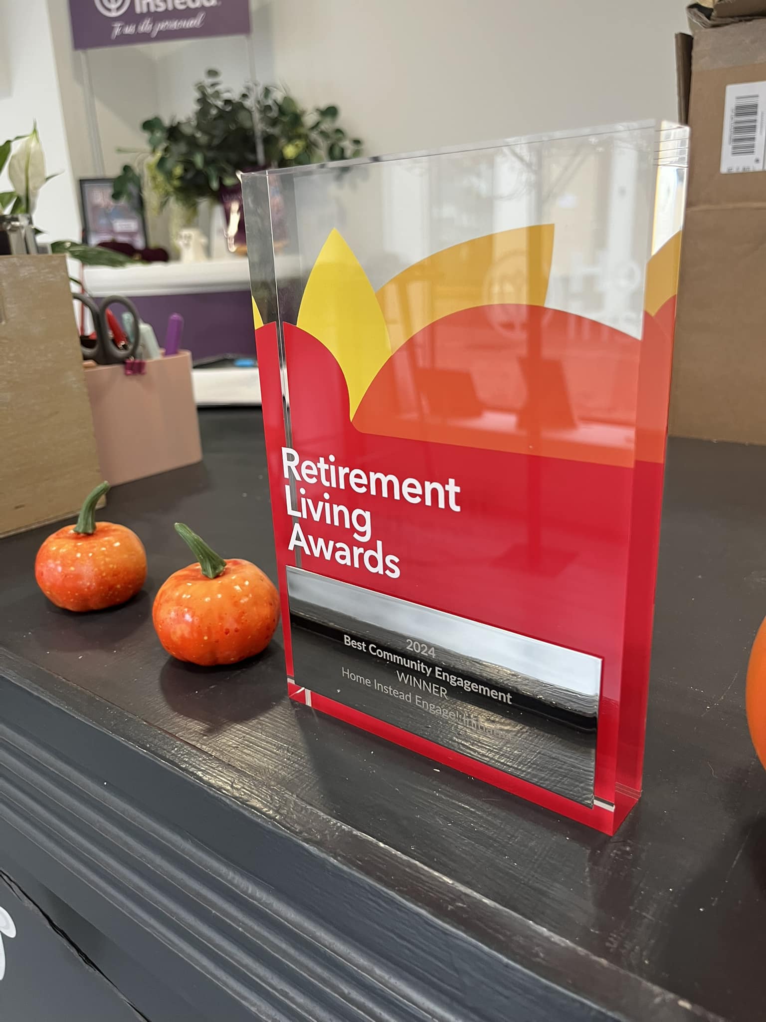 Award plaque for "Best Community Engagement" next to small orange decorative pumpkins on a table. - Home Instead