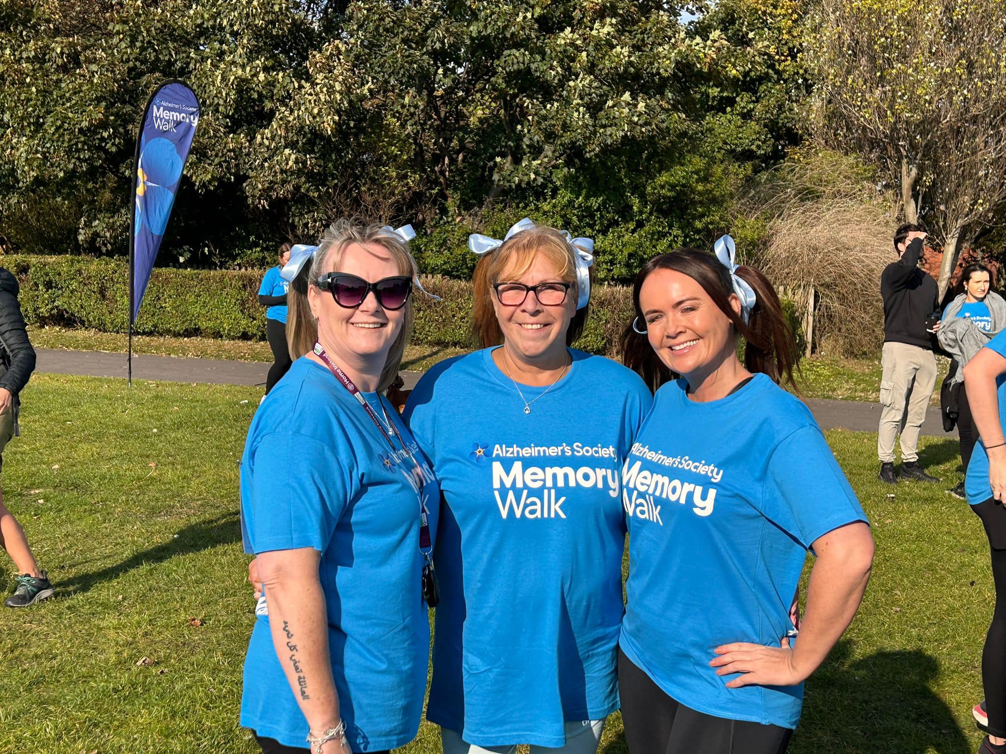 Louise, Debbie and Vicki from the Home Instead team stand side by side at the Alzheimer's society memory walk, dressed in blue t-shirts and blue hair ribbons.