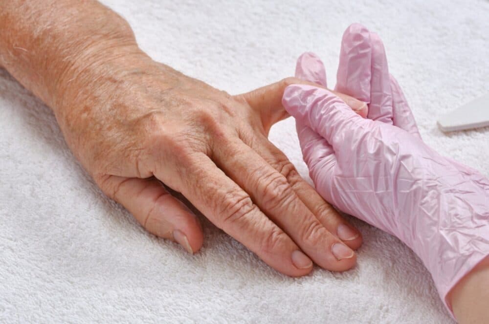 A gloved hand gently holds an elderly person's hand on a white towel, suggesting care or a manicure. - Home Instead