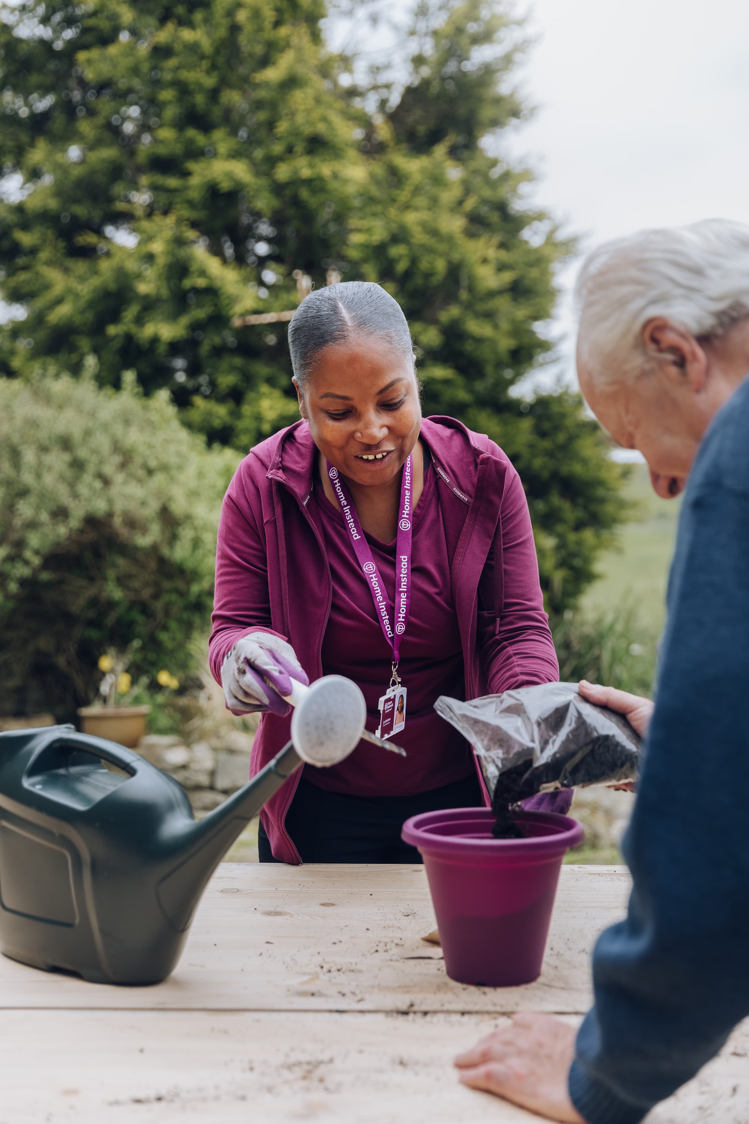 A woman helps an elderly man fill a purple flowerpot with soil outdoors, next to a watering can. - Home Instead