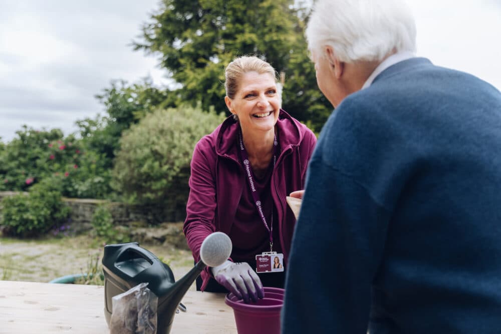 A smiling woman helps an older man with gardening at an outdoor table. - Home Instead