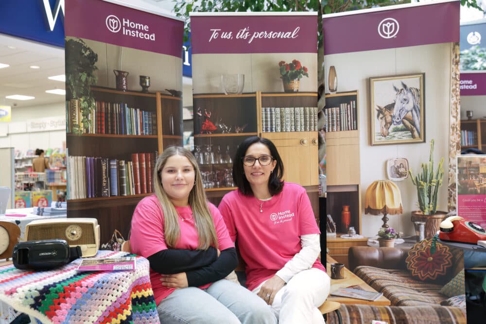 Two smiling women in pink shirts sit at a booth with home decor banners featuring Home Instead branding. - Home Instead