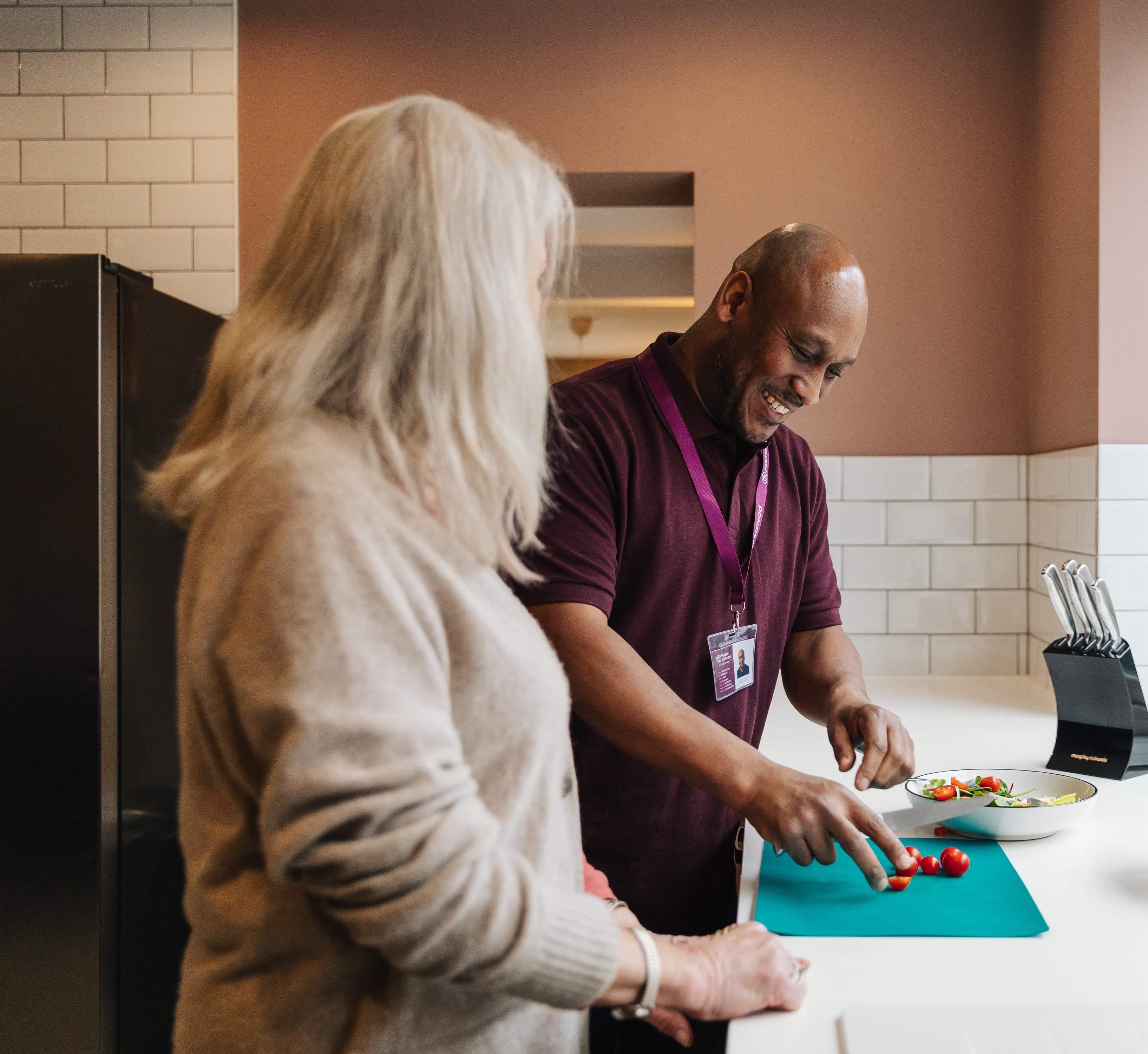 Two people in a kitchen, one smiling man chopping tomatoes on a cutting board, the other watching. - Home Instead
