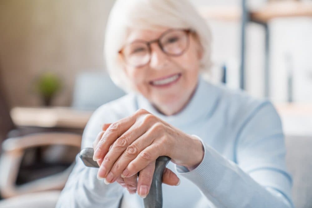 Smiling elderly woman with glasses rests her hands on a cane, sitting indoors. - Home Instead