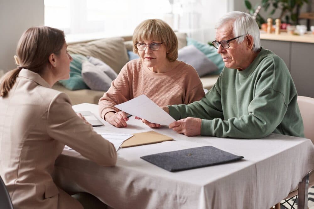 Three people sit at a table, reviewing documents in a well-lit room with a sofa and kitchen in the background. - Home Instead