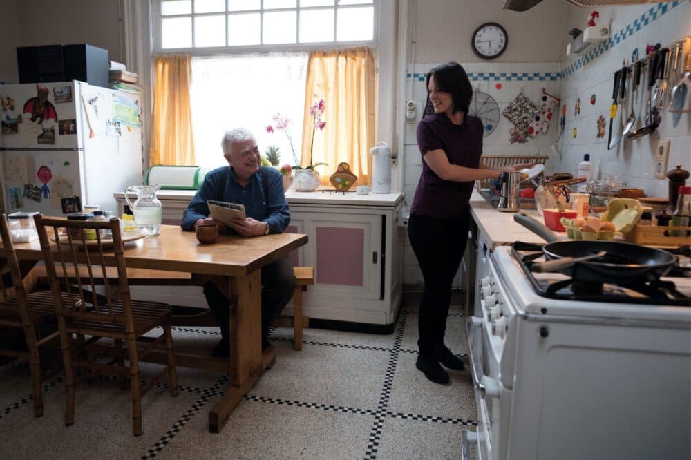An older man sits at a kitchen table while a woman stands nearby, smiling and preparing food. - Home Instead