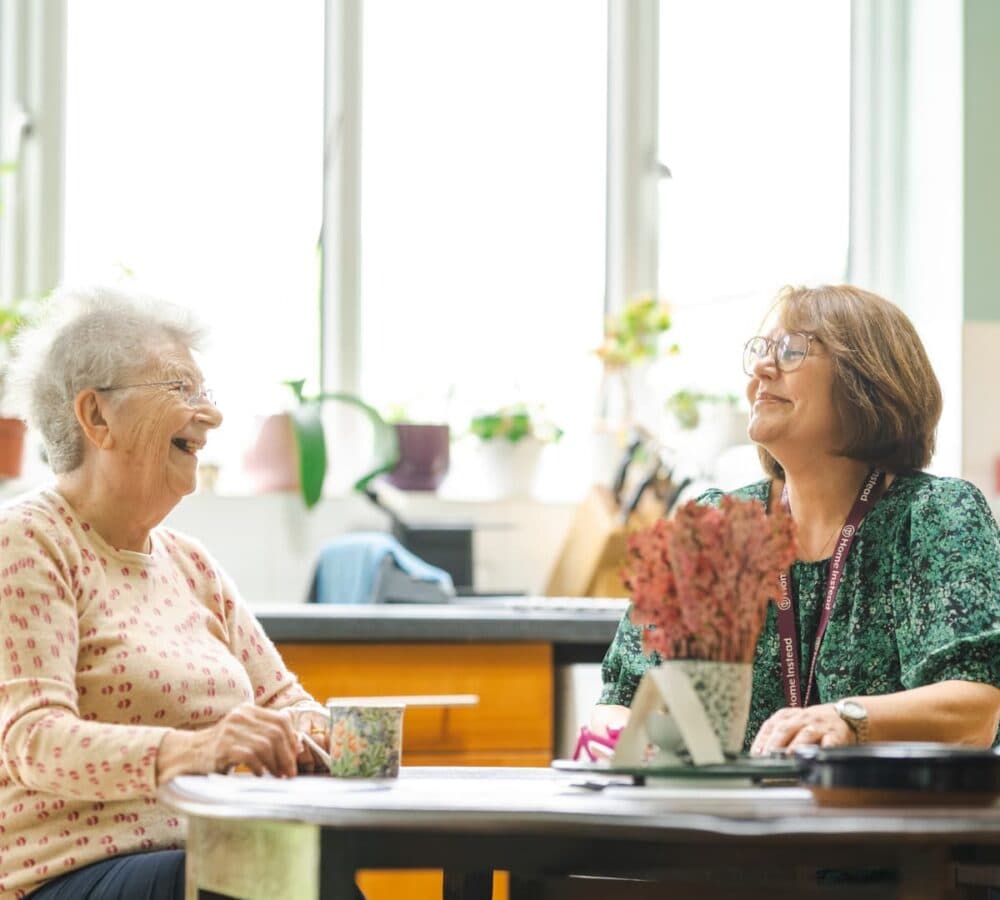 A happy senior wearing eyeglasses sitting inside the kitchen while laughing with her carer wearing green and a pair of eyeglasses with two mugs on top of the table