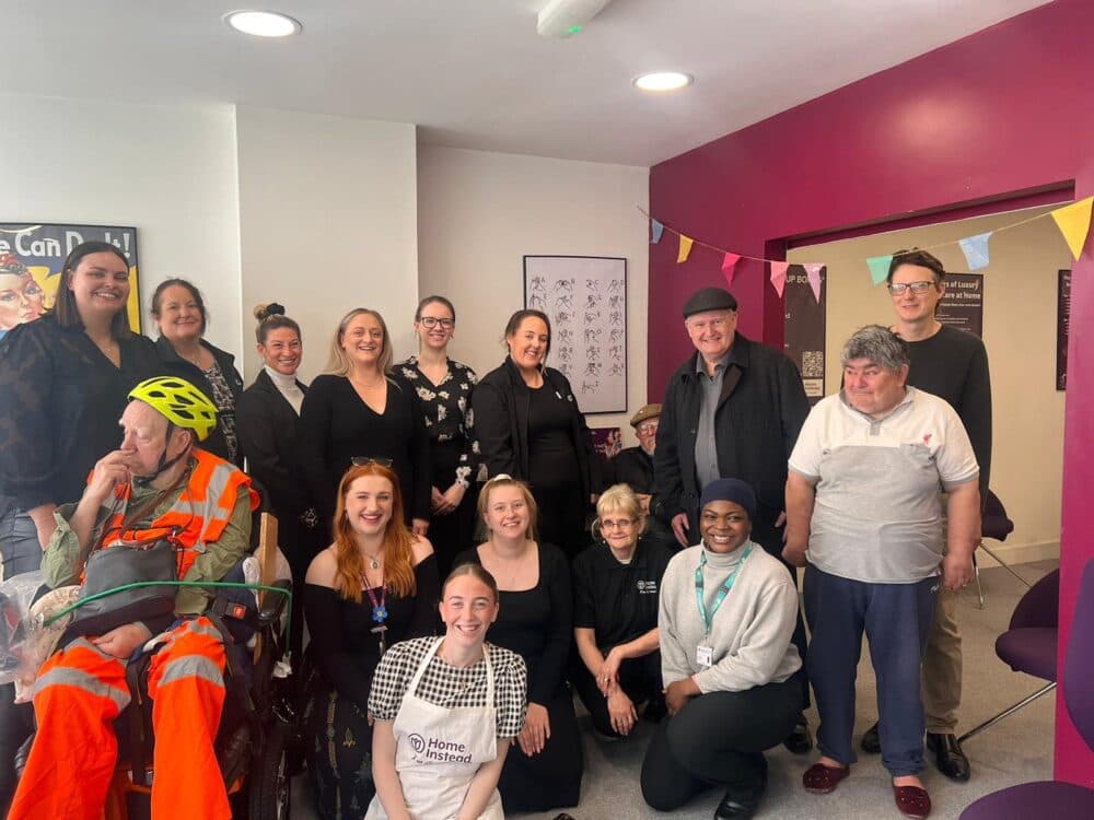 A diverse group of people smiling in a room with colorful walls and bunting. One person is in a reflective uniform. - Home Instead