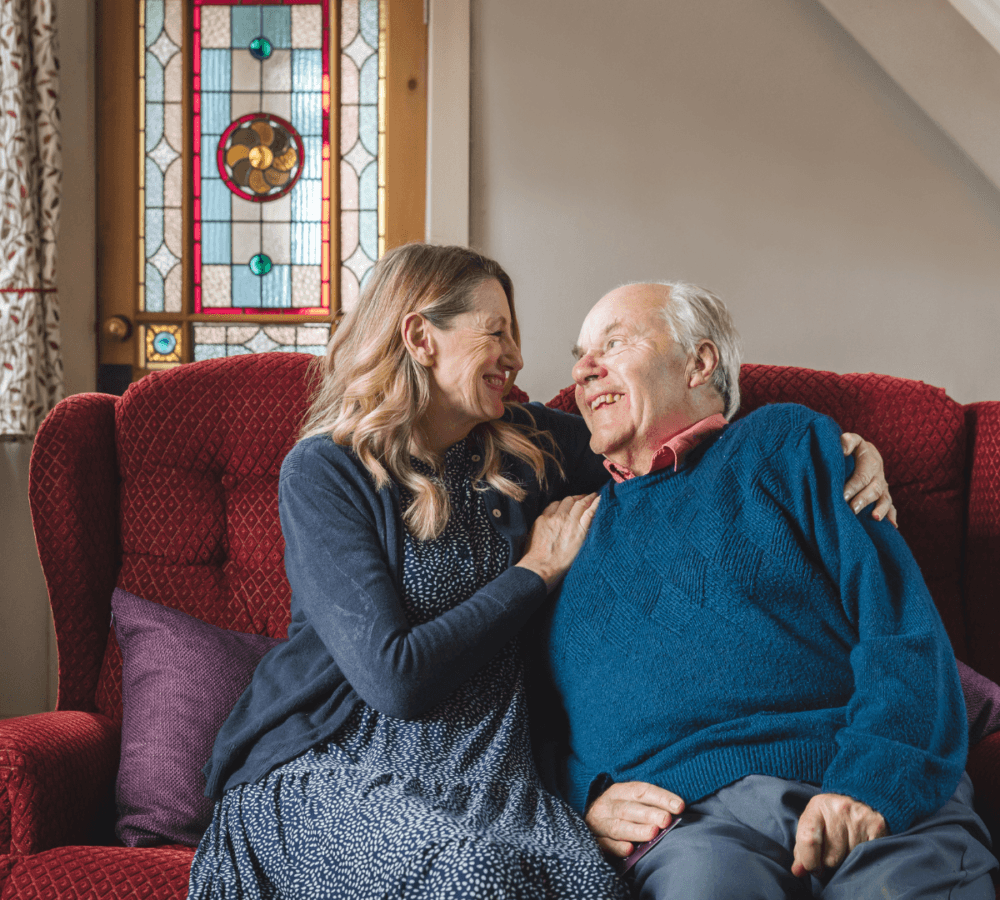 A senior man wearing blue sweater sitting on a couch while smiling and looking at his daughter