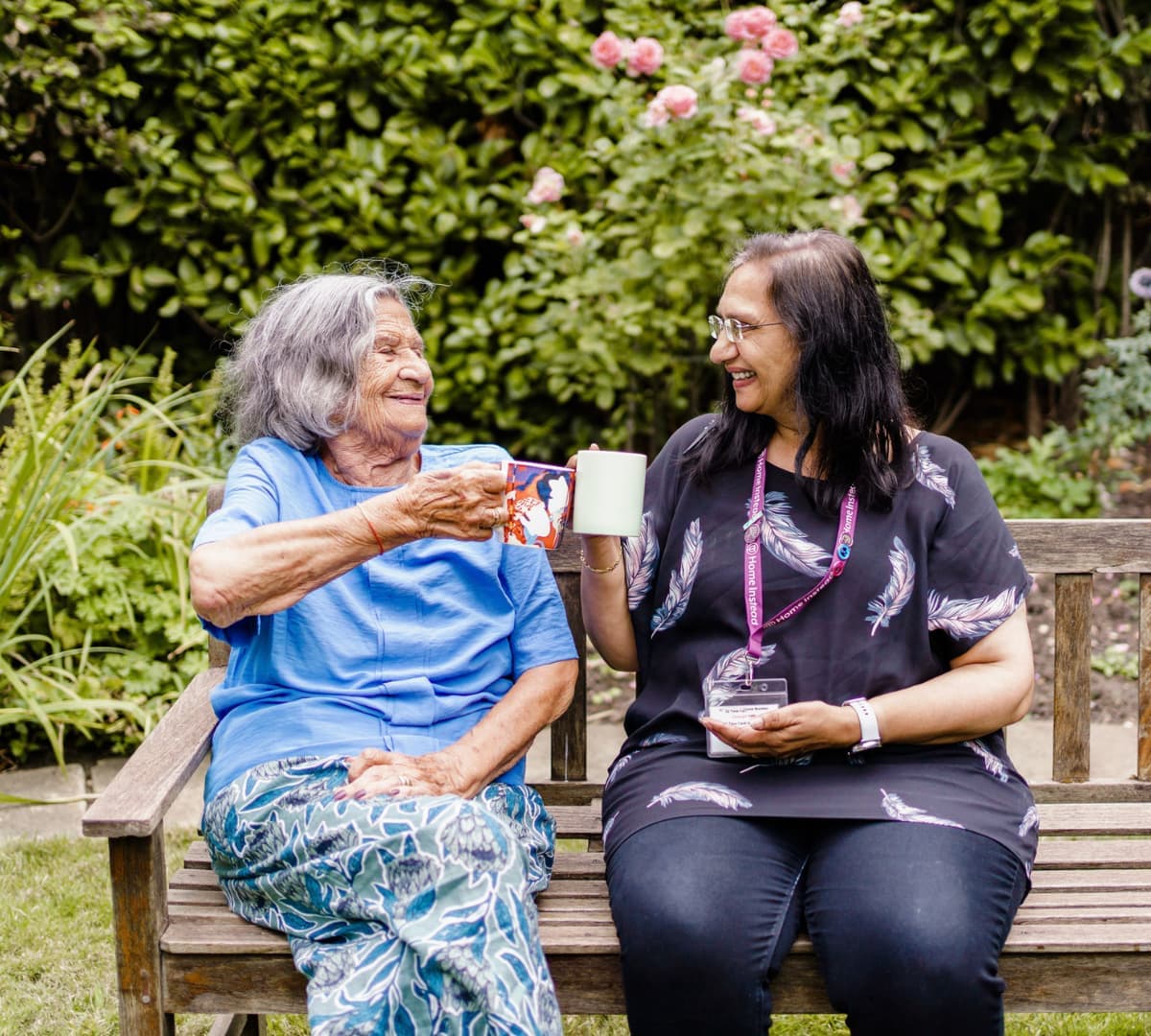 A senior woman with grey hair wearing blue shirt smiling and sitting on a bench while toasting a mug with her carer with long black hair and wearing eyeglasses and black top and pants smiling while in the garden