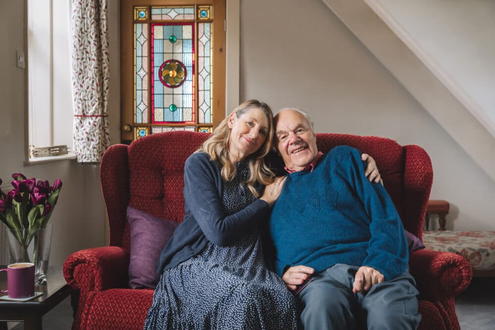 Smiling woman and elderly man sitting together on a red sofa in a cozy, sunlit living room. - Home Instead