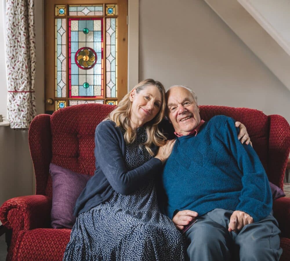 Father and daughter sitting on a couch both happy and smiling while daughter is holding her father