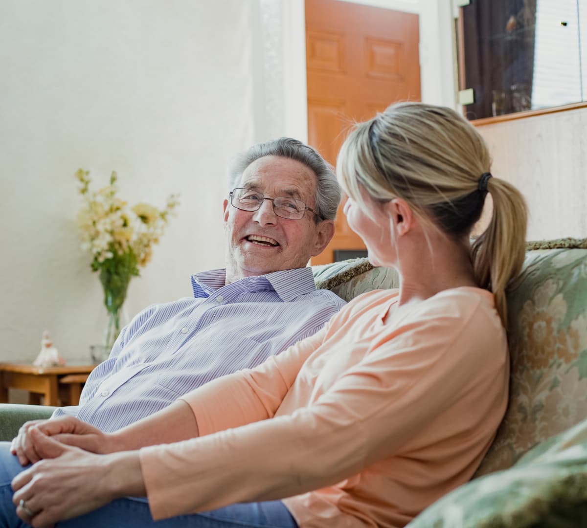 Senior man with daughter sitting on a couch inside a house smiing while looking at each other