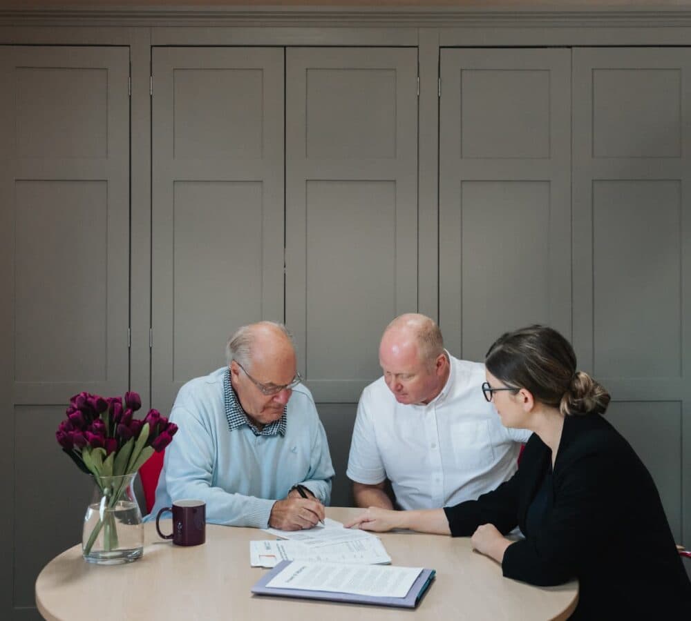 Three people discussing inside a room, two men and 1 woman with papers on the table and a flower vase