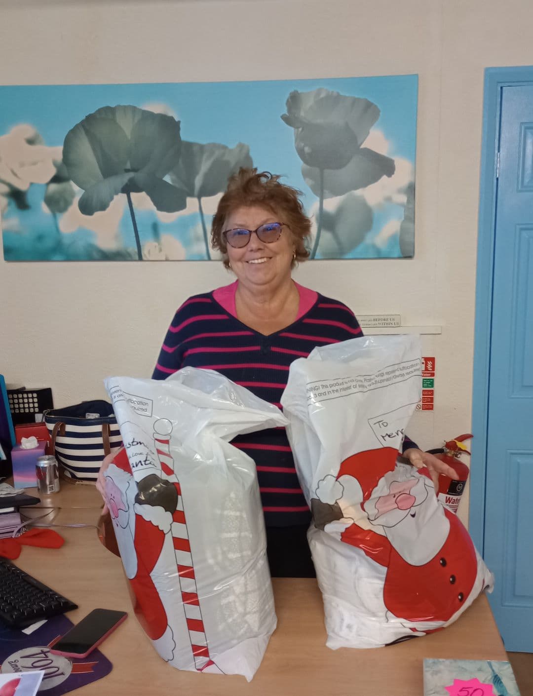 Smiling woman stands behind a desk holding two large festive gift bags with Santa Claus designs. - Home Instead