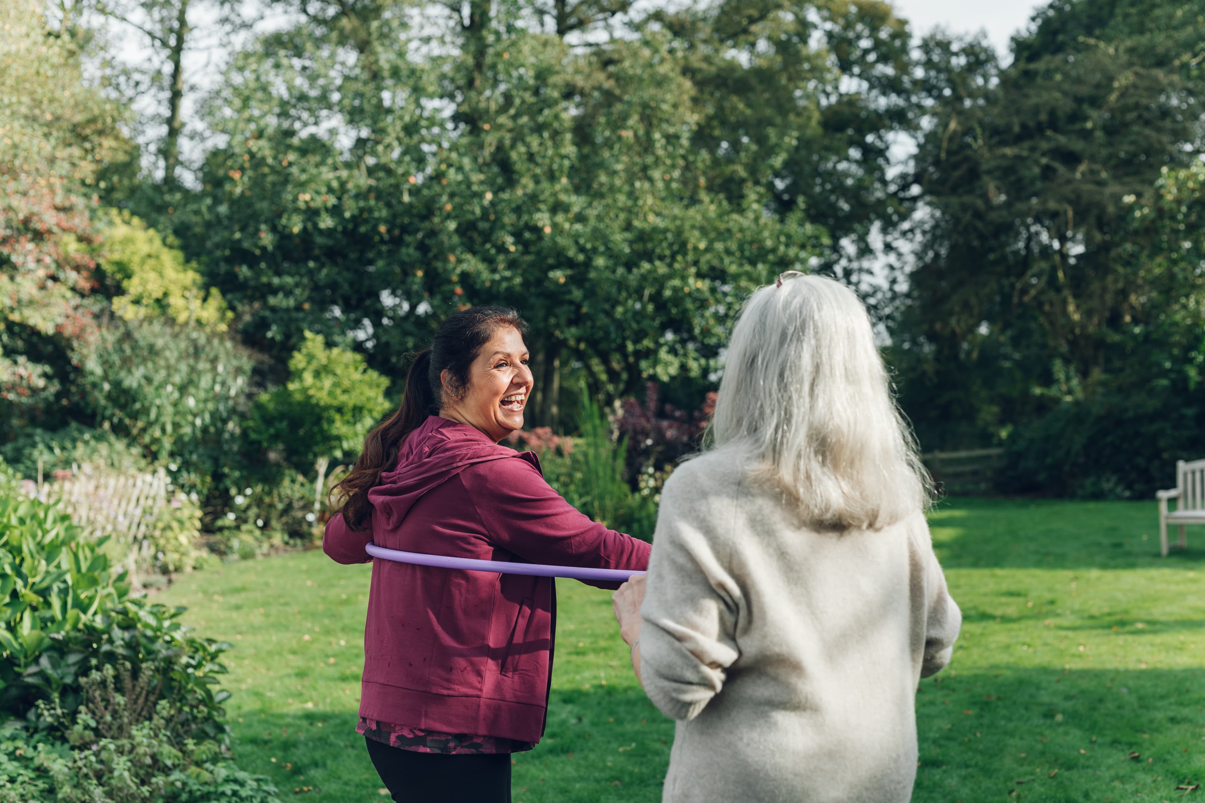 Two women in a garden, one smiling while hula hooping and the other watching her. - Home Instead