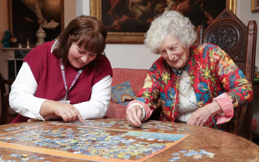 Two women sit at a table smiling and working together on a large jigsaw puzzle. - Home Instead