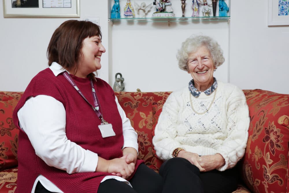 A caregiver and an elderly woman sit smiling together on a patterned red sofa in a cozy room. - Home Instead