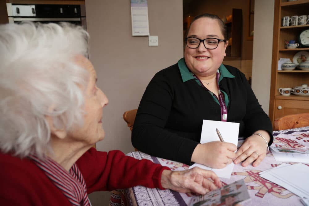 A woman smiles while talking to an elderly lady at a table, holding a notepad and pen. - Home Instead