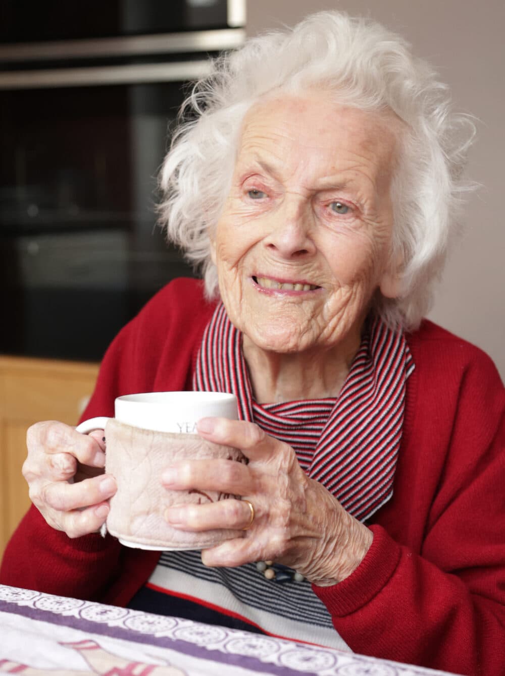 Smiling elderly woman in a red sweater holds a mug while sitting at a table indoors. - Home Instead