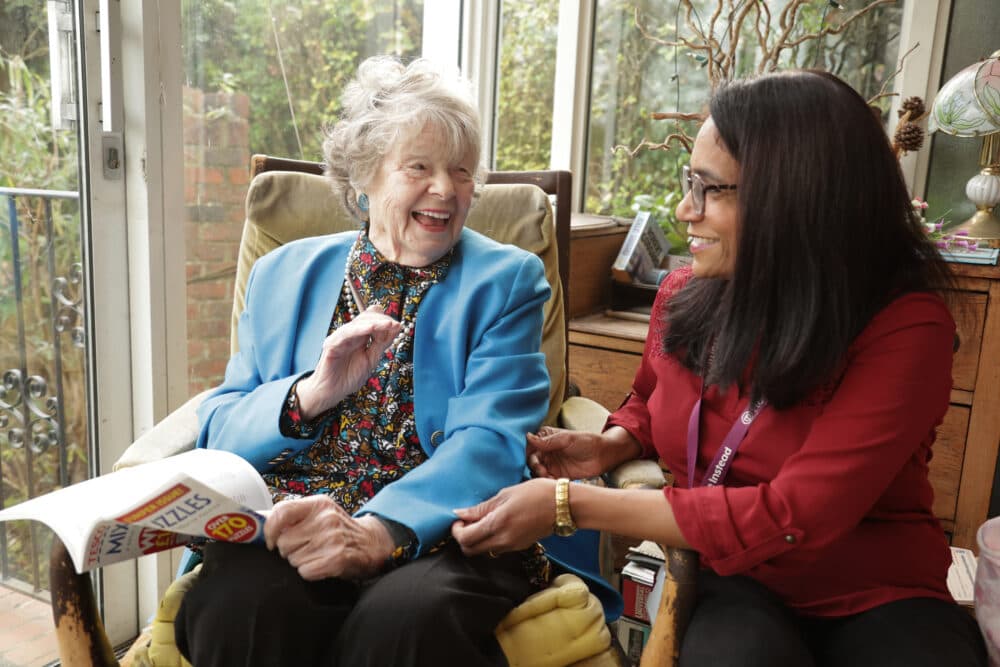 An elderly woman and a younger woman sit together, smiling and talking, in a bright, cozy room. - Home Instead