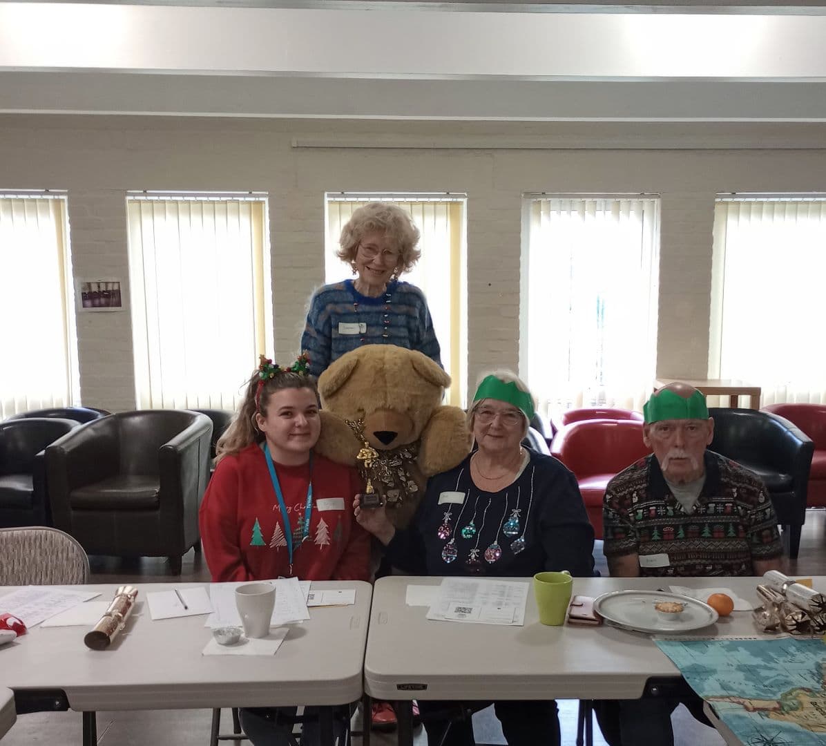 Four people, one holding a large teddy bear, sit and stand at a table decorated for a festive holiday event. - Home Instead