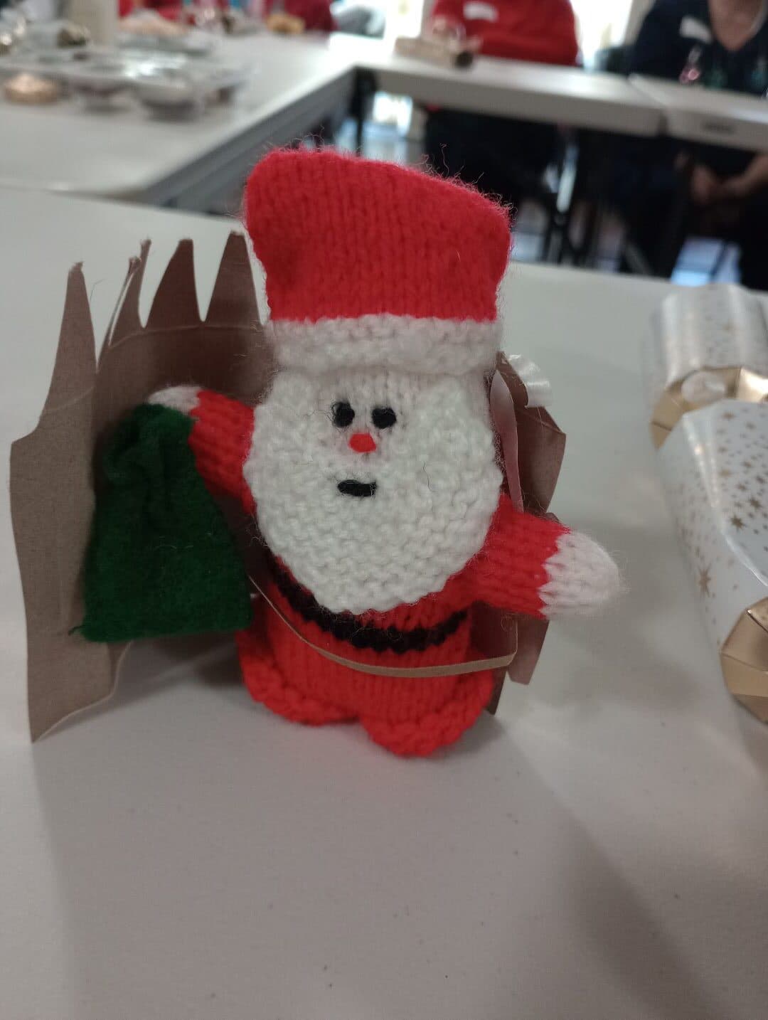 A knitted Santa Claus figurine holding a green sack stands next to a cardboard chimney on a table. - Home Instead