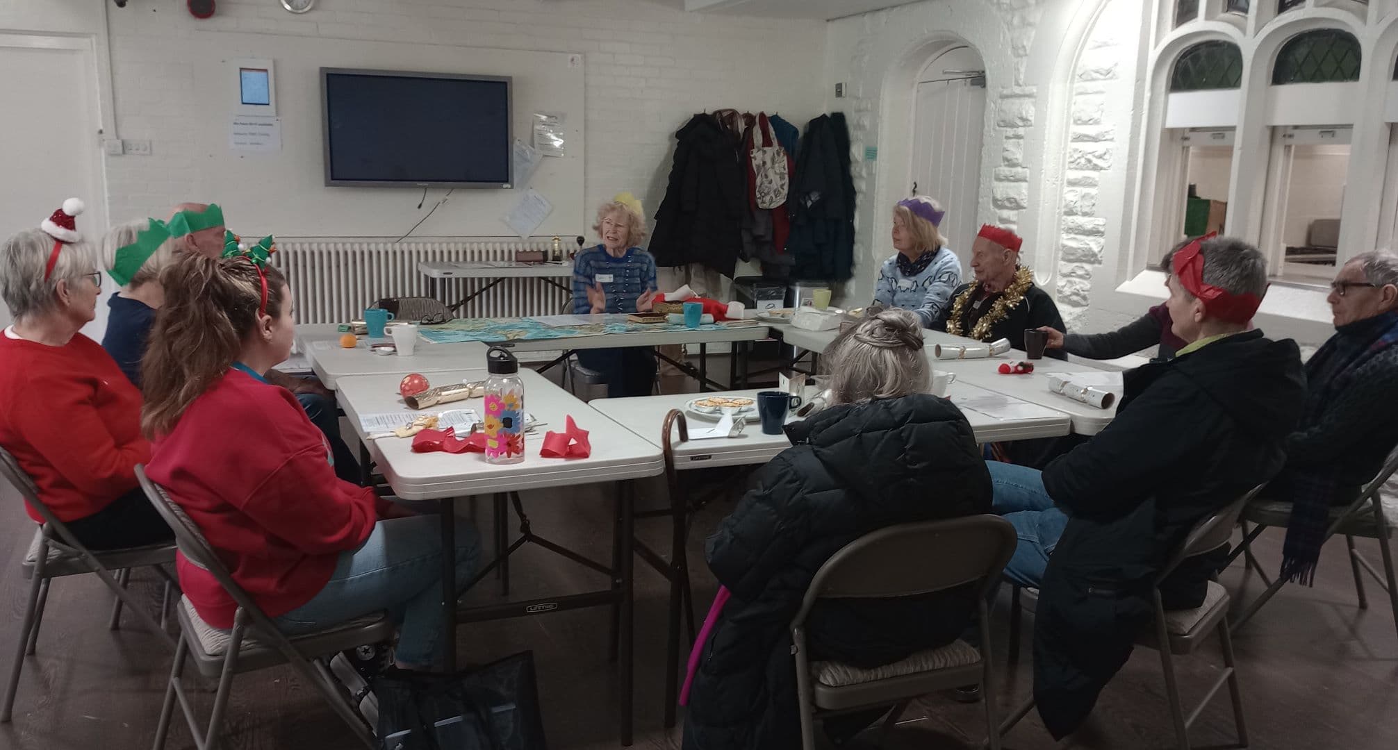 A group of people wearing festive hats sit around tables in a bright room, sharing food and conversation. - Home Instead