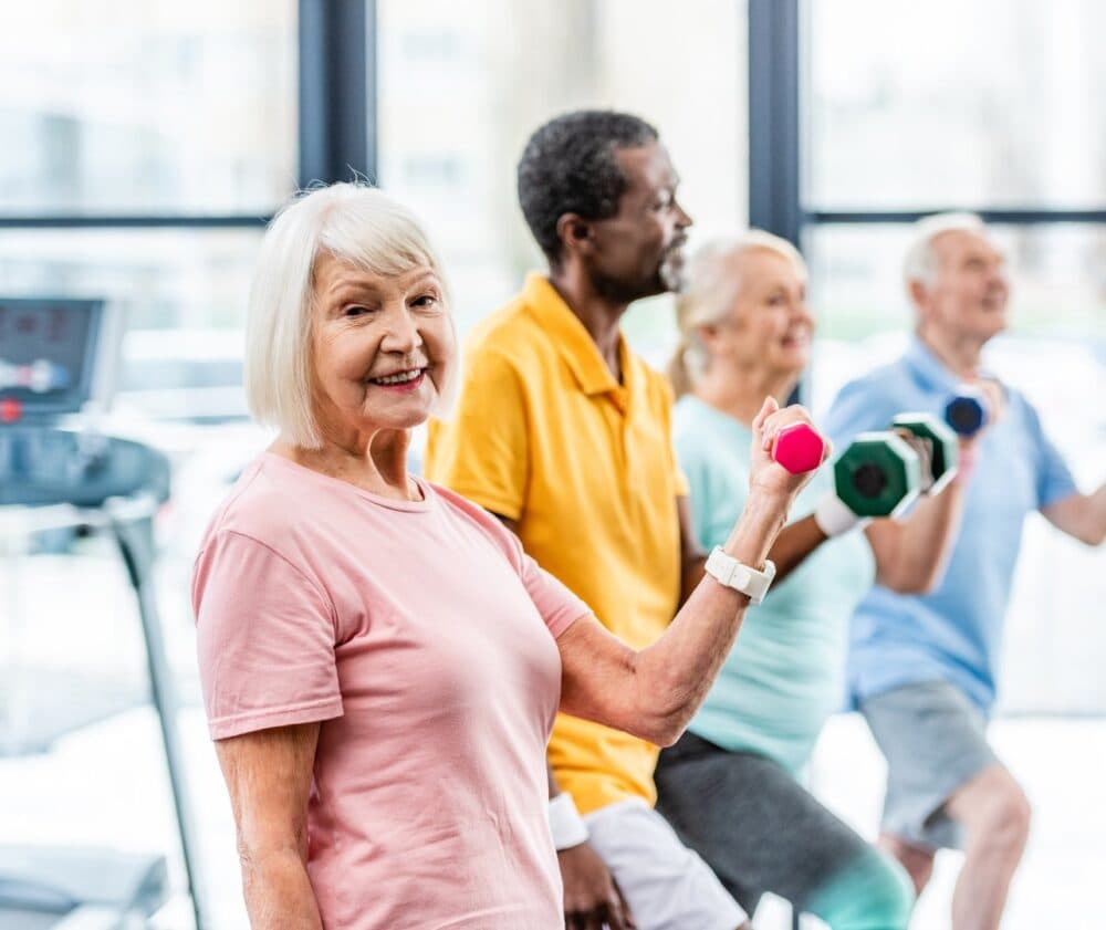 Four smiling seniors exercise with hand weights in a bright gym, with one woman in front looking at the camera. - Home Instead