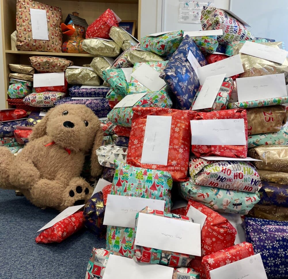 A large teddy bear sits by a pile of wrapped gifts and envelopes on a carpeted floor. - Home Instead