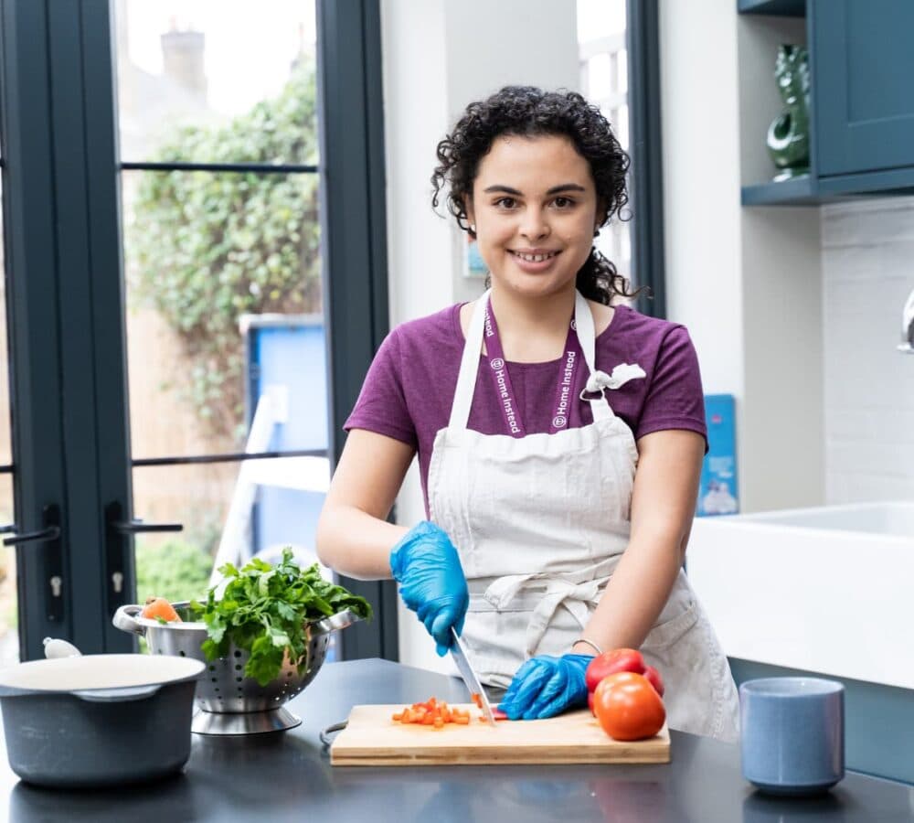 Woman with curly hair in the kitchen wearing apron and gloves while cutting some vegetables