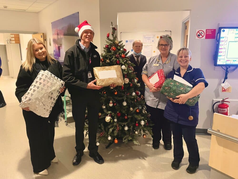 Five people stand by a Christmas tree in a hospital, holding wrapped gifts and smiling at the camera. - Home Instead