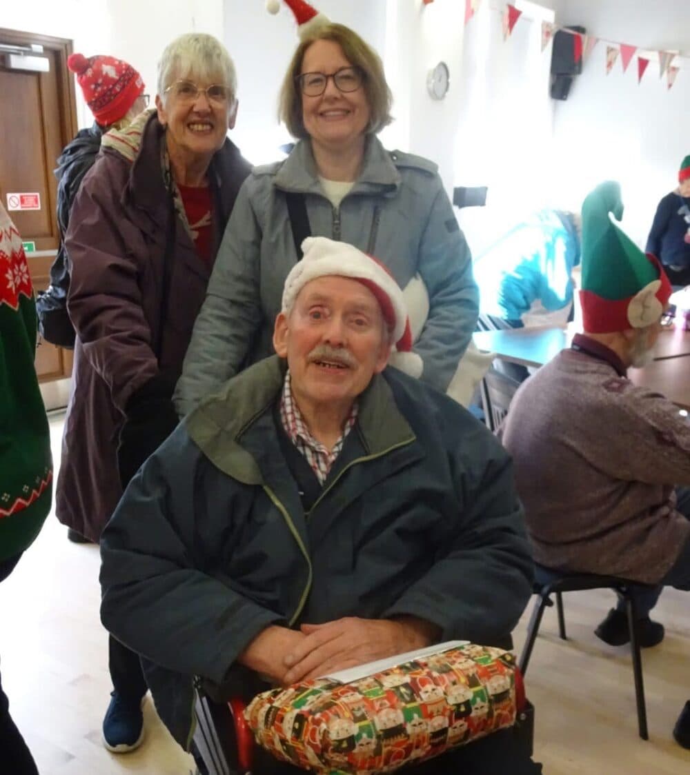 Three smiling older adults in festive attire, one in a Santa hat and wheelchair, at a holiday gathering indoors. - Home Instead