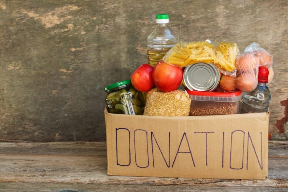 A cardboard box labeled "DONATION" filled with food items like pasta, apples, and canned goods. - Home Instead
