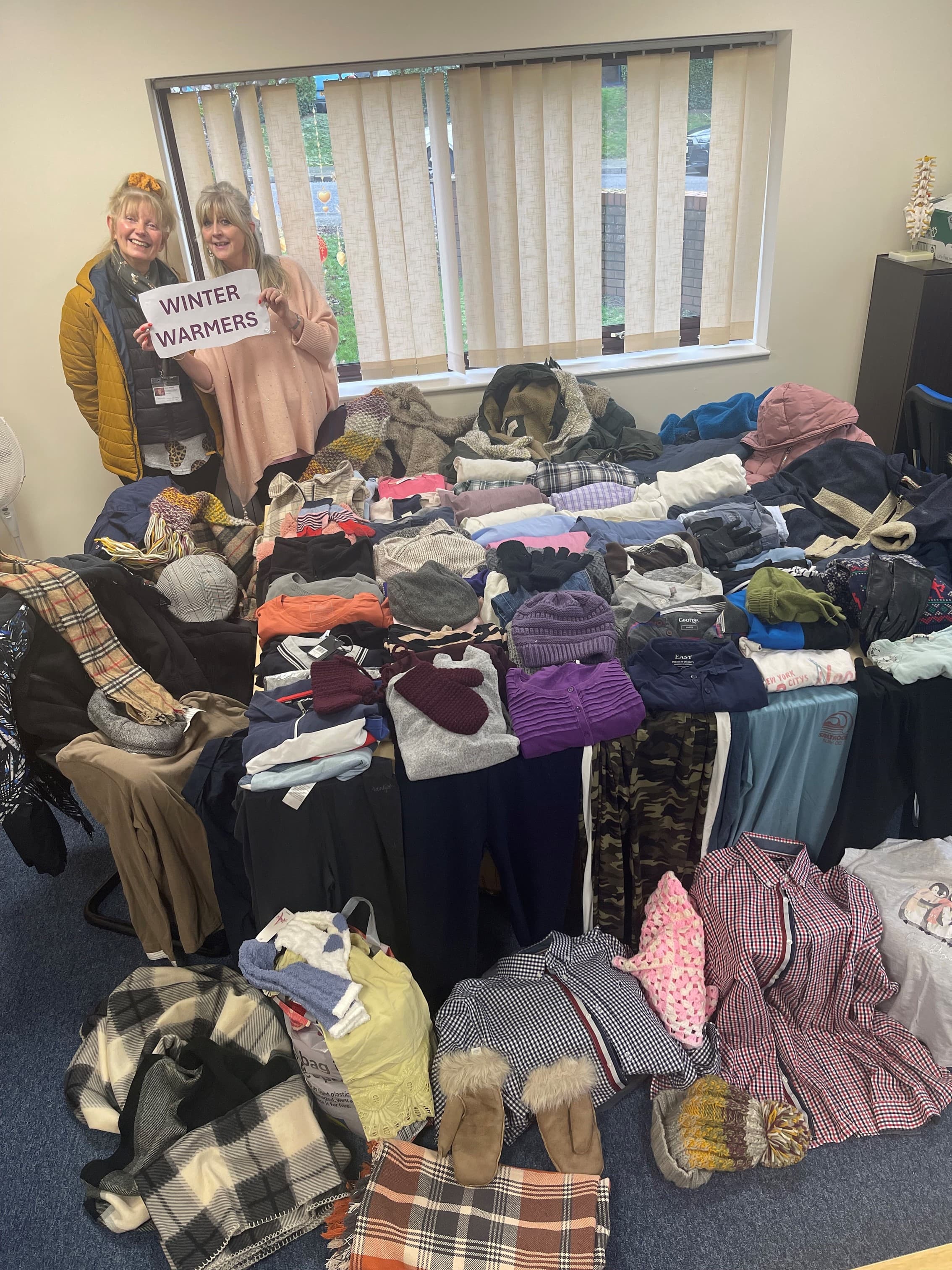 Two people stand by a table full of winter clothing, holding a "Winter Warmers" sign in an office. - Home Instead