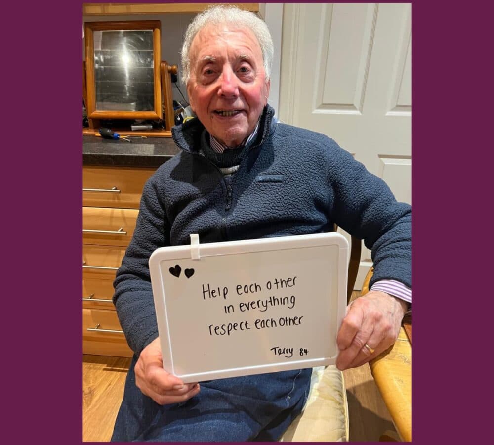Older man with white hait and sitting on a chair inside the home holding a sign that says Help each other in everything, respect each other.