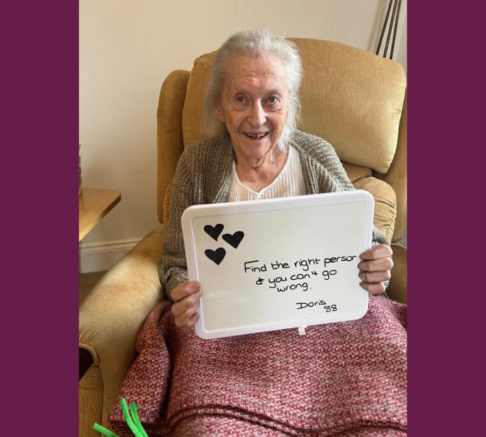 Older woman with grey hair sitting on a beige couch wearing sweater and holding a sign board