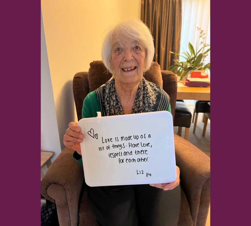 Older woman Liz with white hair sitting on a couch while holding a sign that says Love is made up of lots of things. Have love, respect and be there for each other.