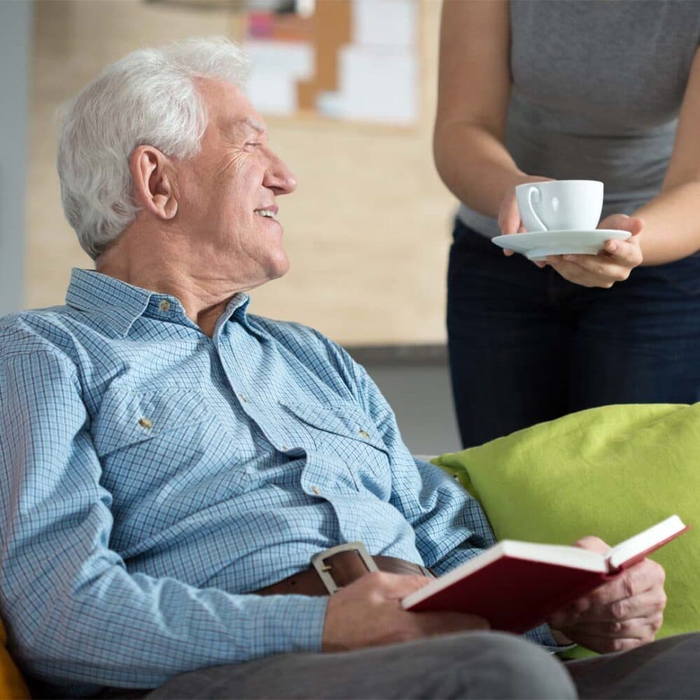 Care professional offering elderly man a cup of tea at home