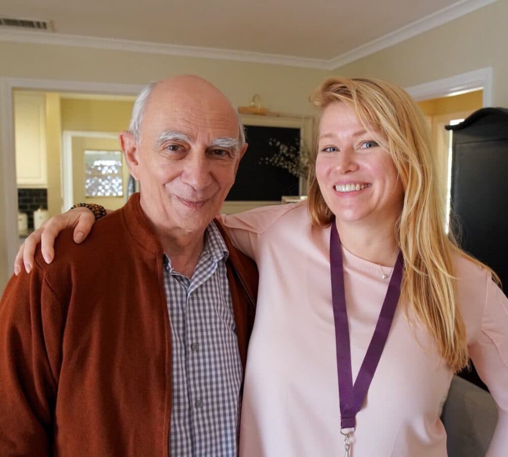 Older man with little grey hair smiling and swearing a sweater together with his young female carer with long blonde hair both happy inside the home