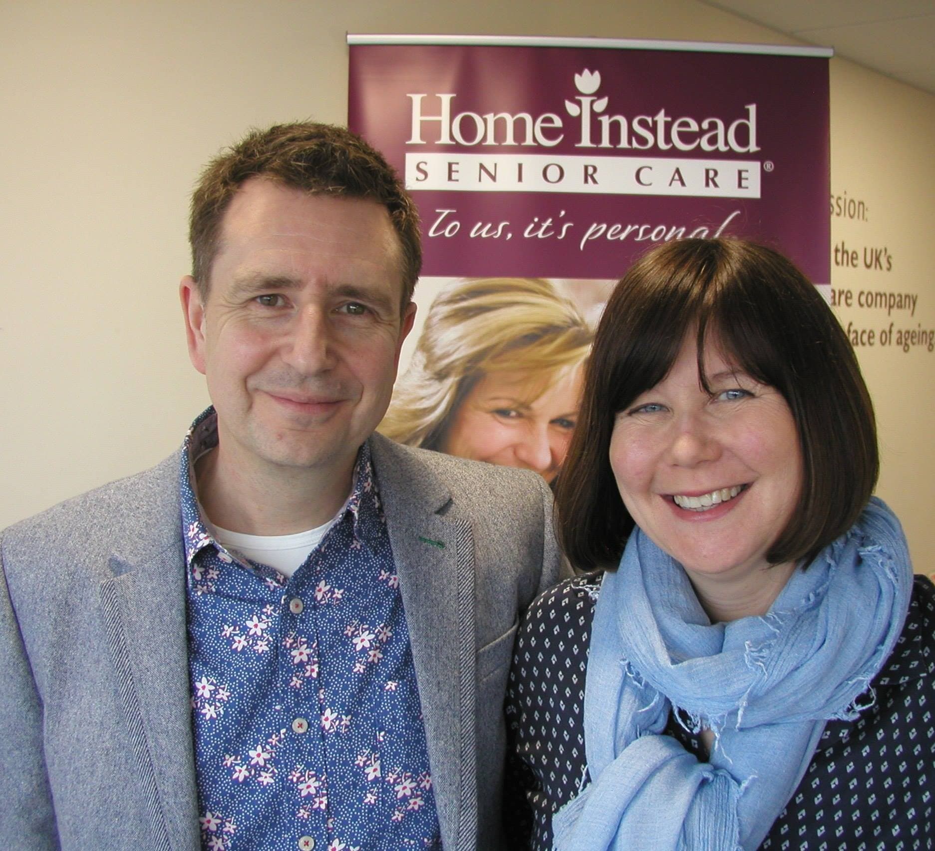 Two smiling people stand in front of a Home Instead Senior Care sign, posing for a photo indoors. - Home Instead